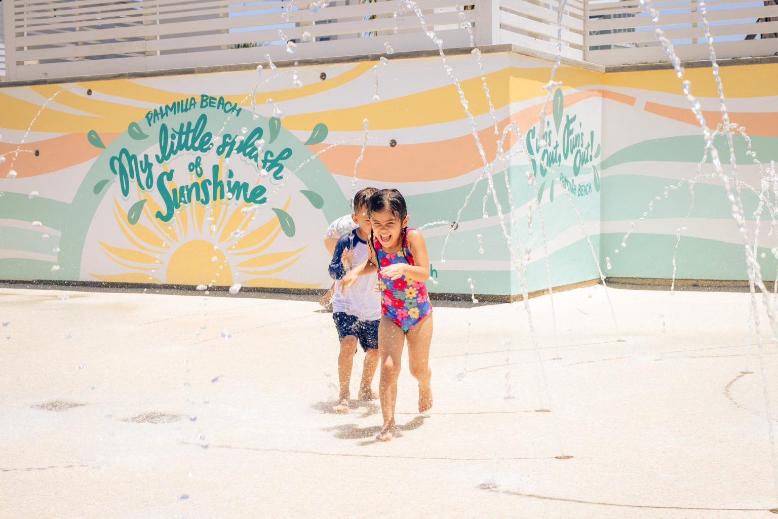 Children running through the splash pad at The Oasis, beside the painted sunshine mural