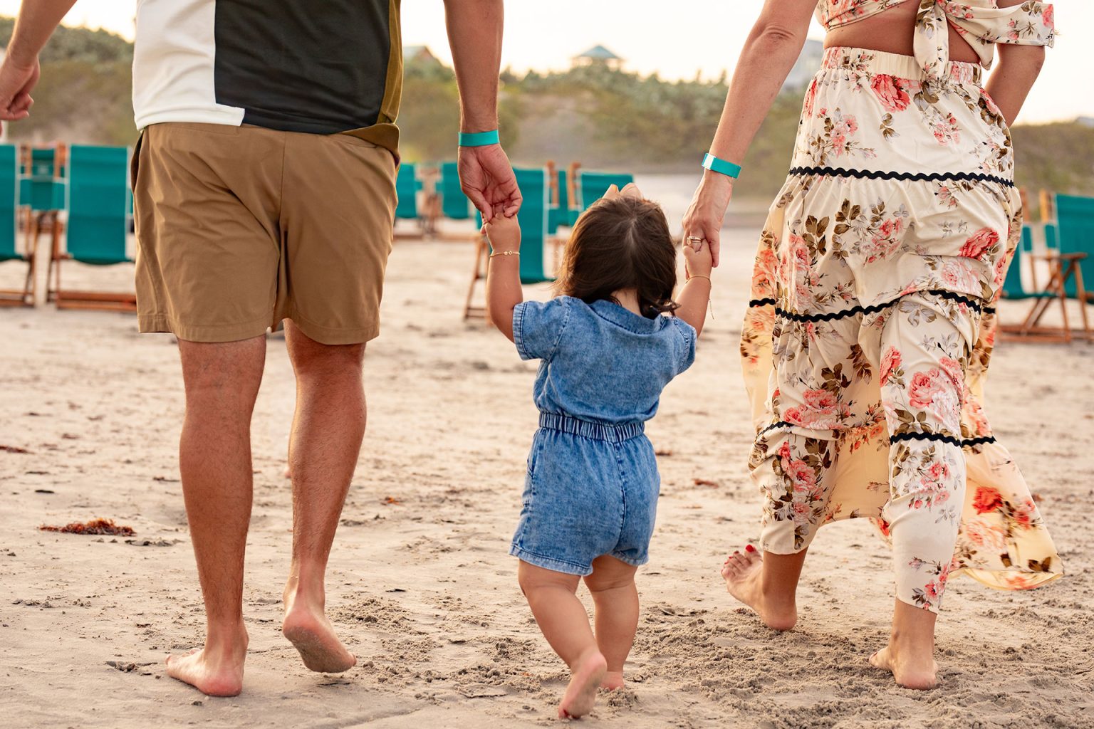 Parents walking a toddler across the sand toward teal beach chairs