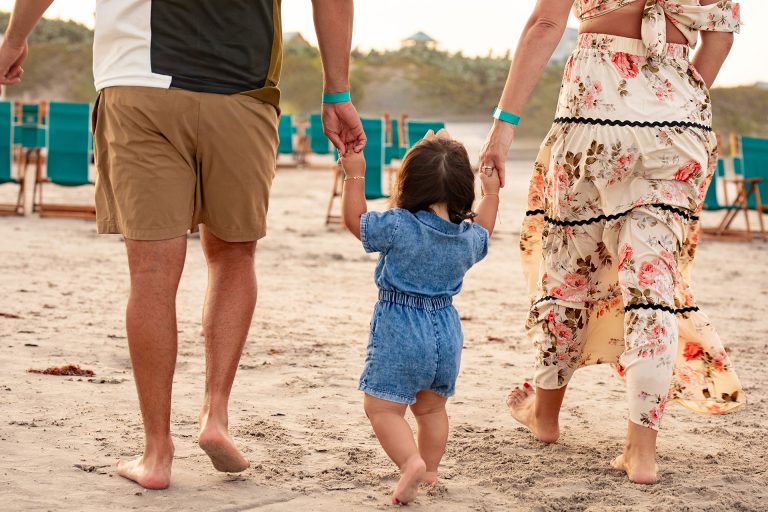 Parents walking a toddler across the sand toward teal beach chairs