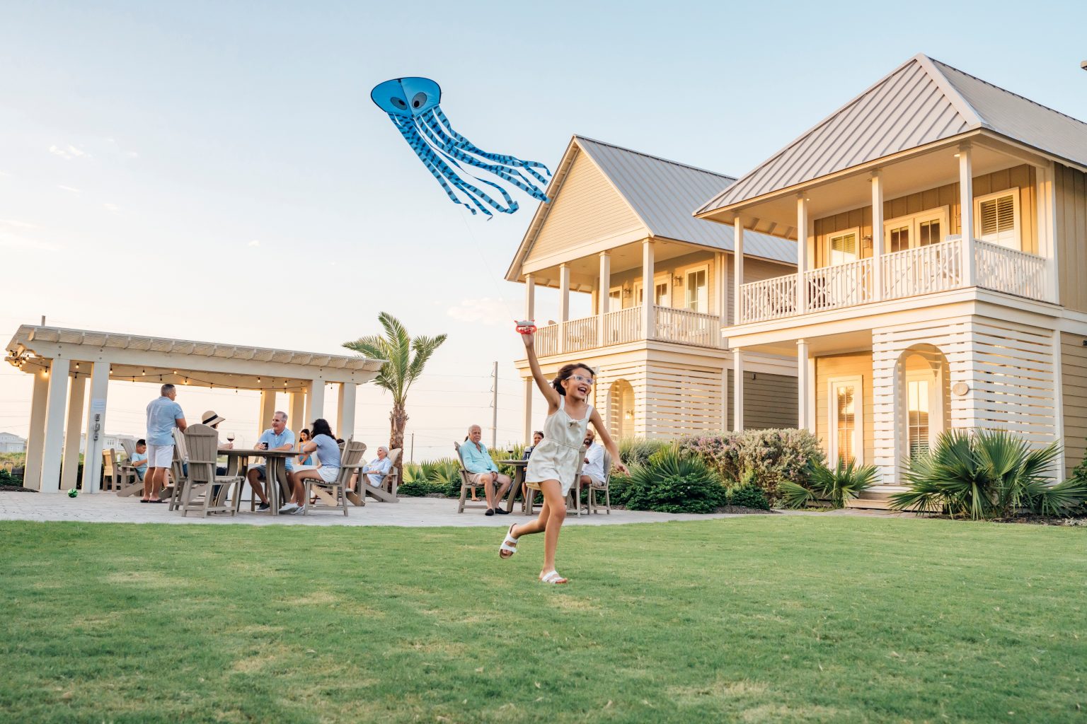 A girl flying a kite on the lawn beside Palmilla Beach homes while families gather under a pergola