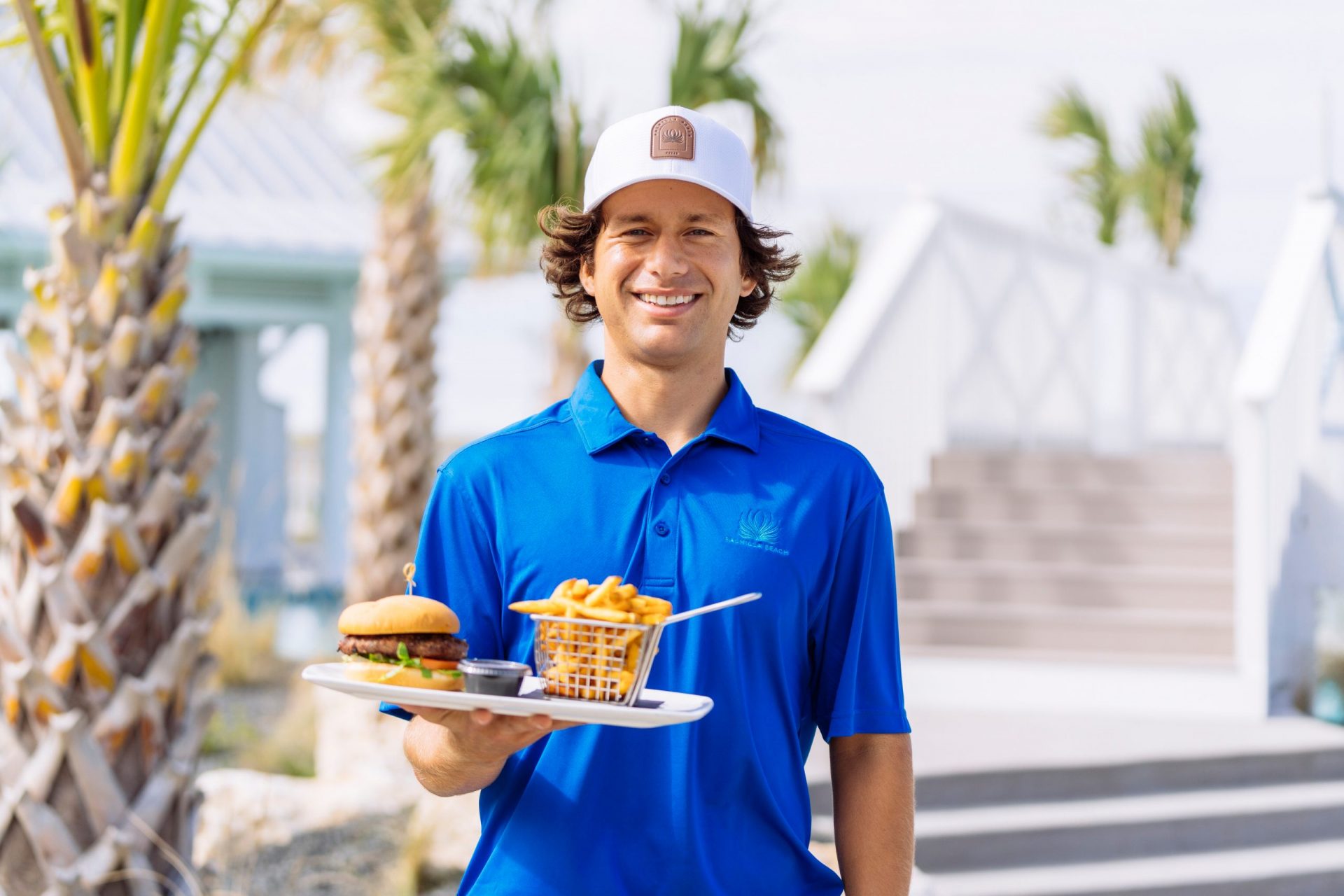 Palmilla Beach server carrying a burger and fries poolside beneath the palms